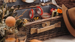 Een picknickopstelling met een rieten mand die een fles, glazen en bestek bevat, een bord met cupcakes en bessen, en een boterham op een deken.