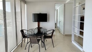 Ein geräumiges Zimmer mit Deckenventilator, einem Glastisch mit Stühlen, einem Fernseher und einem großen Fenster mit durchsichtigen Vorhängen.