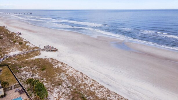 Een strand met een steiger die zich uitstrekt over de oceaan, een zandgebied met weinig begroeiing en een zwembadgedeelte met een hek.