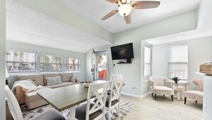 A spacious living room with a large dining table, comfortable seating, a ceiling fan, and a TV mounted on the wall.