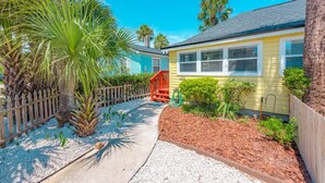 A pathway leading to a yellow house with a red staircase, surrounded by palm trees and a wooden fence.