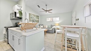 A compact kitchen with a central island, a dining area with a table and chairs, a sofa, and a ceiling fan. - Pink Dolphin 3| Beachside Cottage| Walk Everywhere (Jacksonville Beach)