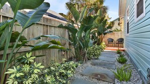 Un sendero en un jardín que conduce a una terraza de madera con sillas, rodeada de exuberante vegetación tropical y una cerca de madera.