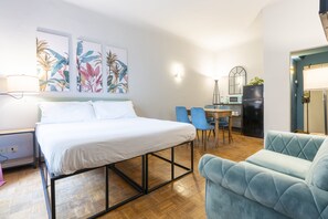 A modern hotel room with a bed, a desk with a chair, a blue armchair, and a kitchen area. - Panzani 13 - Studio Apartment, Sleeps 2 (Firenze)
