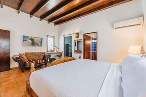 A bedroom with a large bed, a desk with a chair, a mirror, and an air conditioning unit.