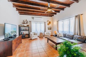 A spacious living room with a wooden ceiling, a flat-screen TV, a bookshelf, two white armchairs, a leather sofa, and a coffee table.