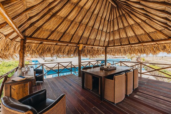 A covered outdoor seating area with a thatched roof, wooden flooring, and a pool in the background.