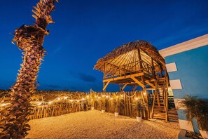 A thatched-roof structure with a wooden frame, surrounded by a gravel area with potted plants and string lights.