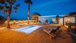 A pool area with lounge chairs, a thatched-roof structure, and a blue building. - Villa Rancho Azul Mins to Palm Beach & Eagle Beach (Noord)