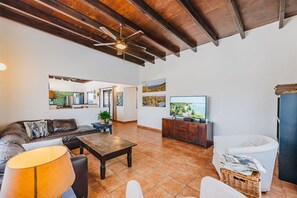 A spacious living room with a sofa, coffee table, and TV, a dining area with a table and chairs, and a kitchen visible through an open door.