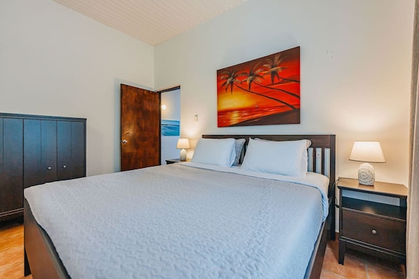 A neatly made bed with a blue blanket, a wooden wardrobe, a nightstand with a lamp, and a painting of palm trees on the wall.