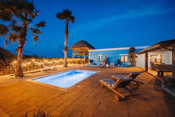 A pool area with lounge chairs, a thatched-roof structure, and a blue building.