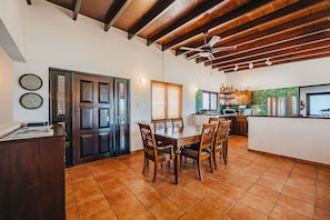 A dining area with a wooden table and chairs, a kitchen with a green backsplash, and a ceiling fan.