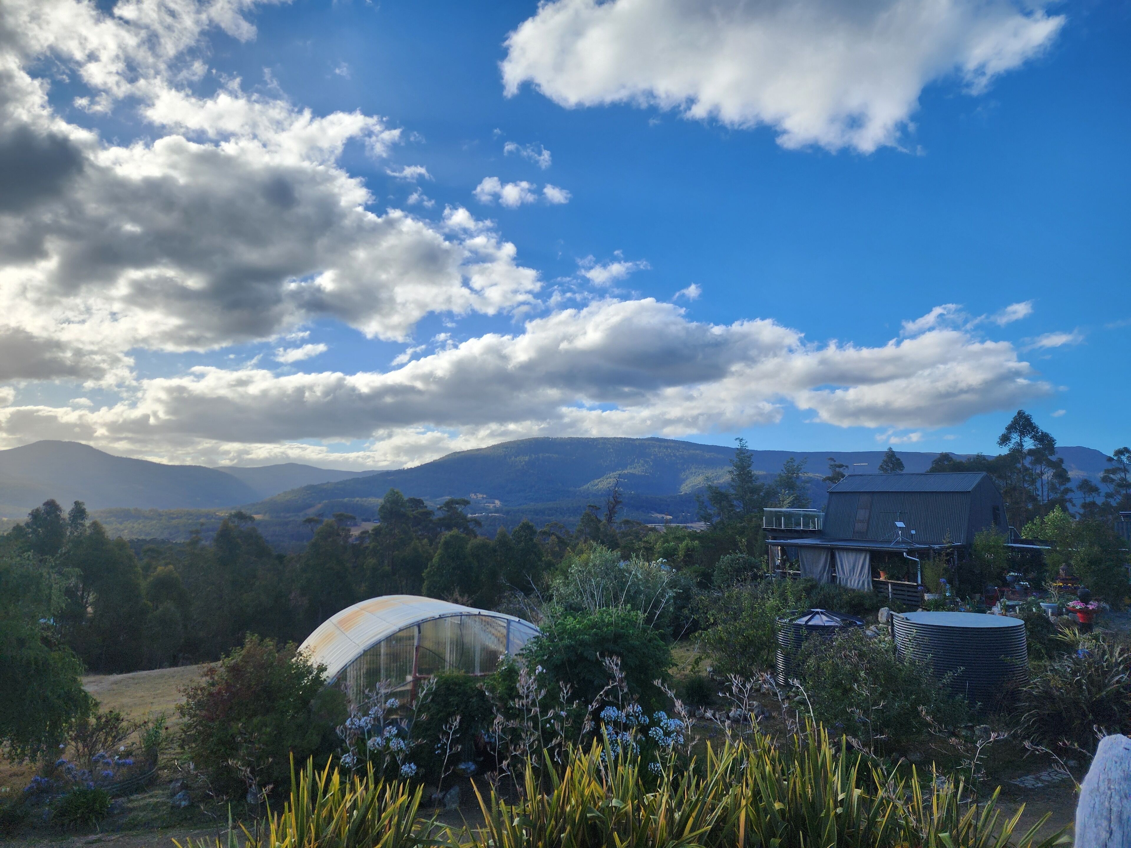 A greenhouse in a garden with a house in the background and mountains in the distance.