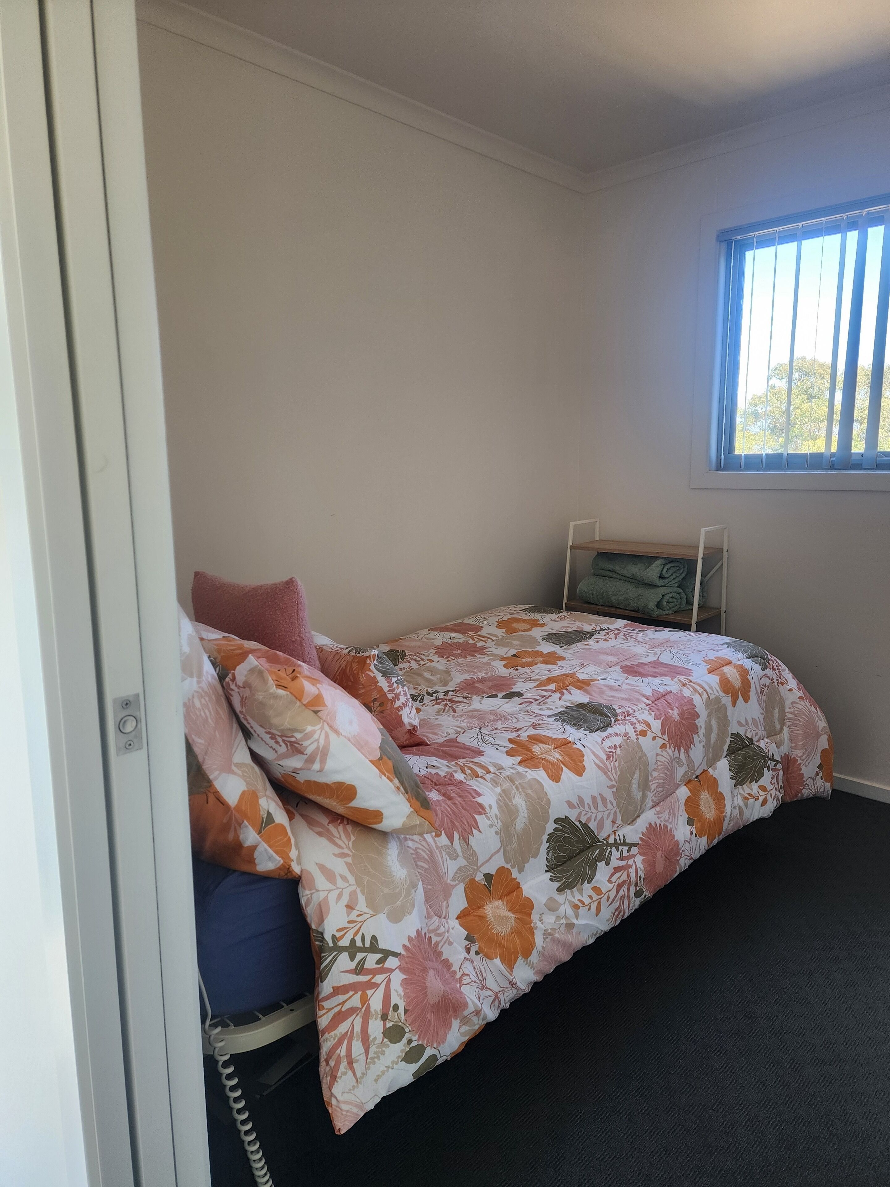 Une petite chambre avec un lit simple, une couvre-lit à motifs floraux, une fenêtre avec des stores et une table de chevet avec une lampe.