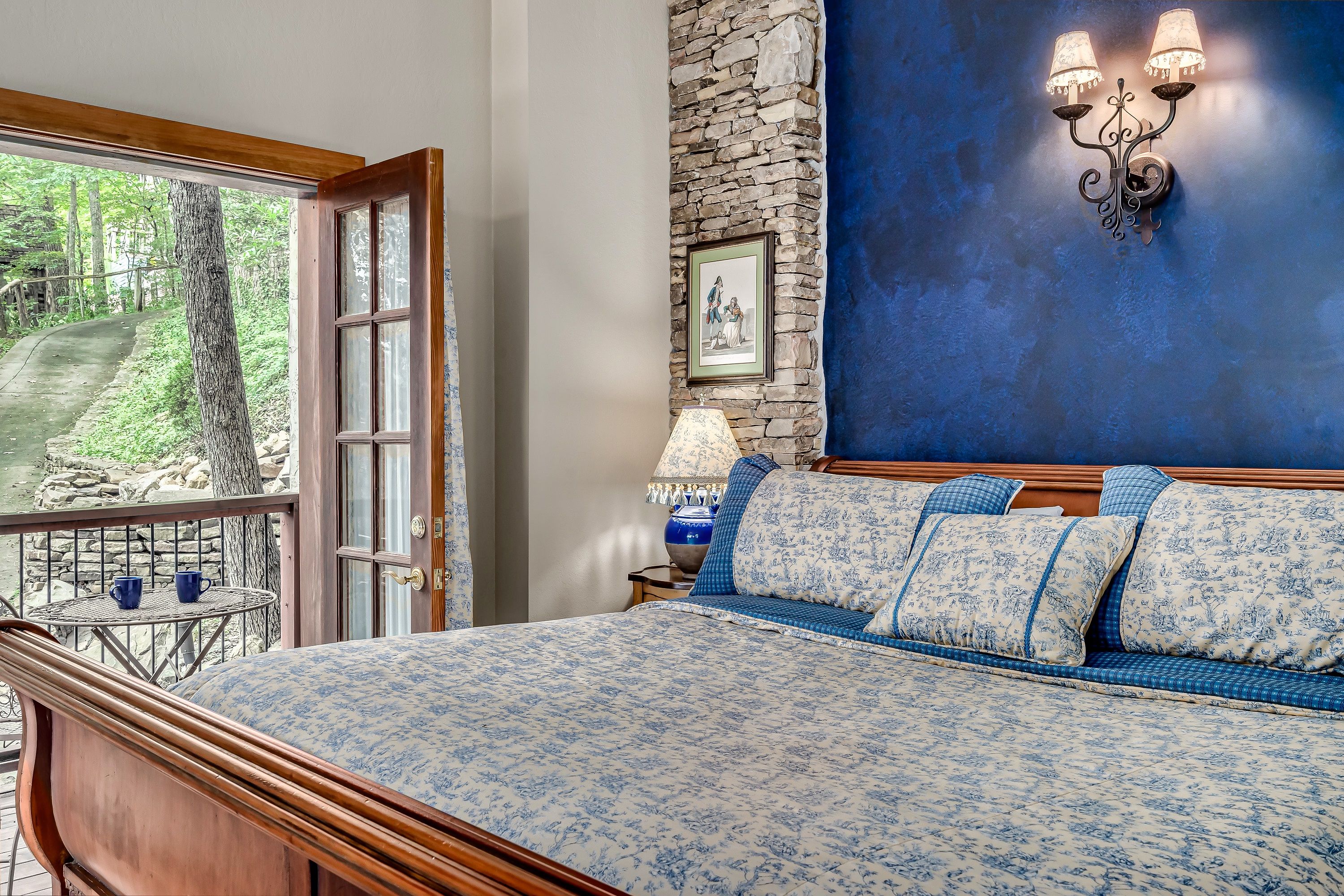 A bedroom with a bed, a stone wall, a picture frame, a nightstand, a lamp, and a view of greenery outside.