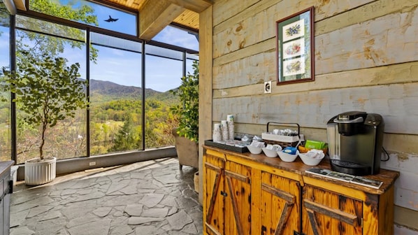 A rustic wooden cabin with a stone floor, a coffee station with a Keurig machine, and a view of a mountainous landscape through large windows.