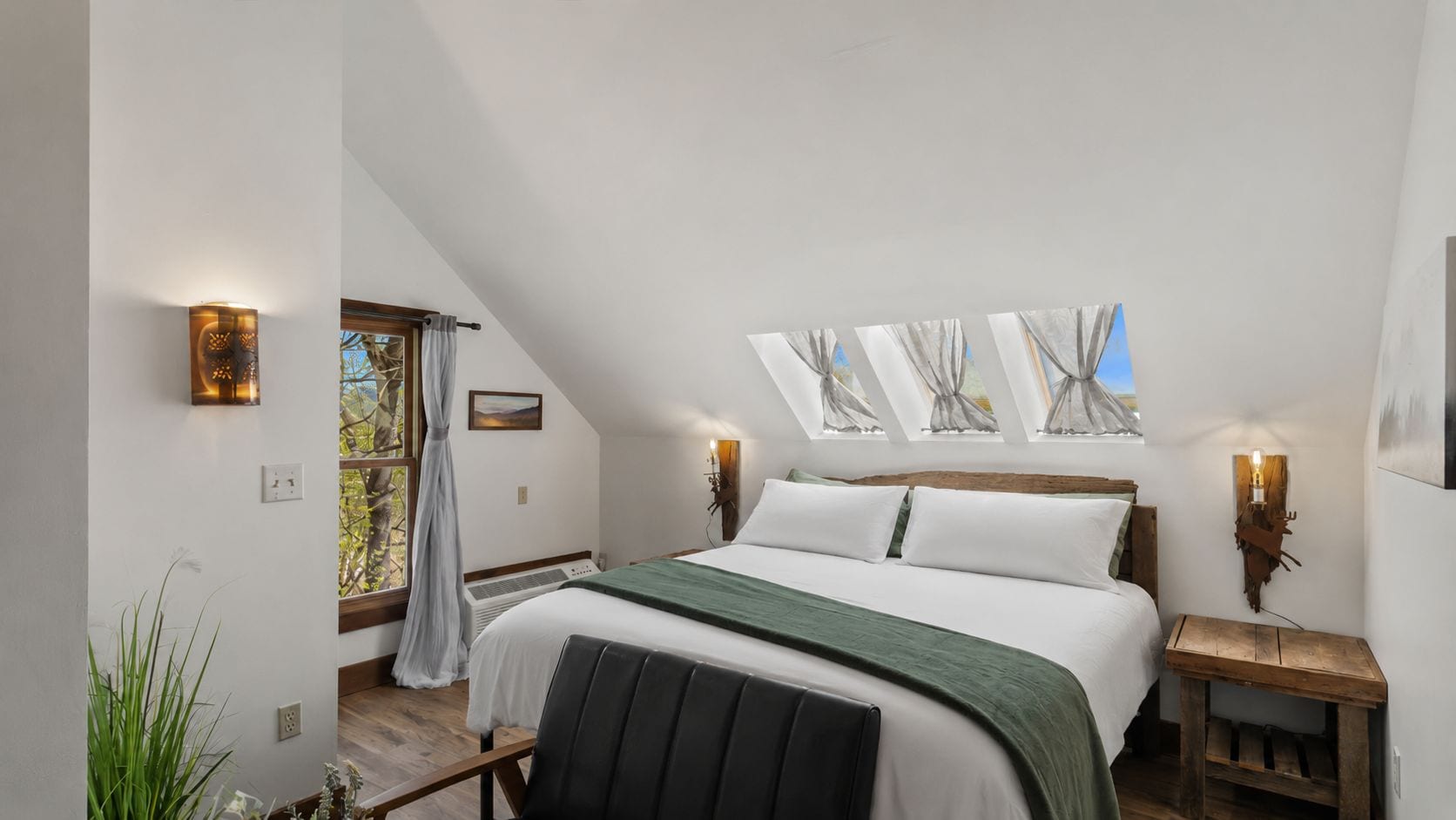 Een slaapkamer met een schuine kap, een bed met een groene sprei, twee nachtlampjes, een raam met gordijnen en een klein houten tafeltje.