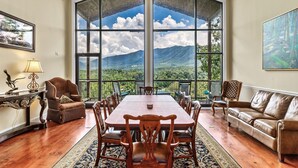 Un'ampia sala da pranzo con un grande tavolo e sedie, un divano, e vista sulle montagne attraverso ampie finestre.