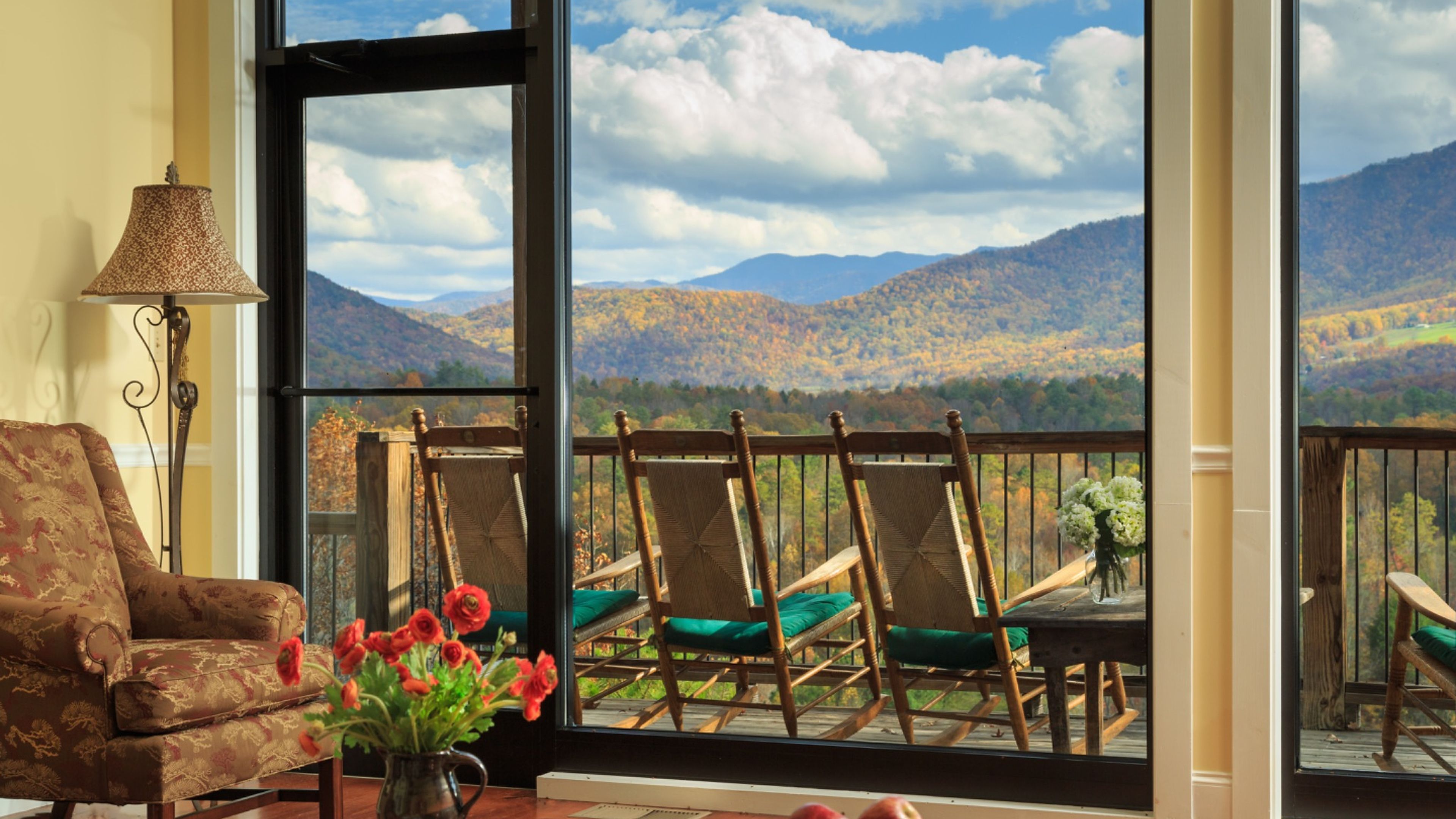 A room with a view of mountains, a chair, a table, and a lamp.