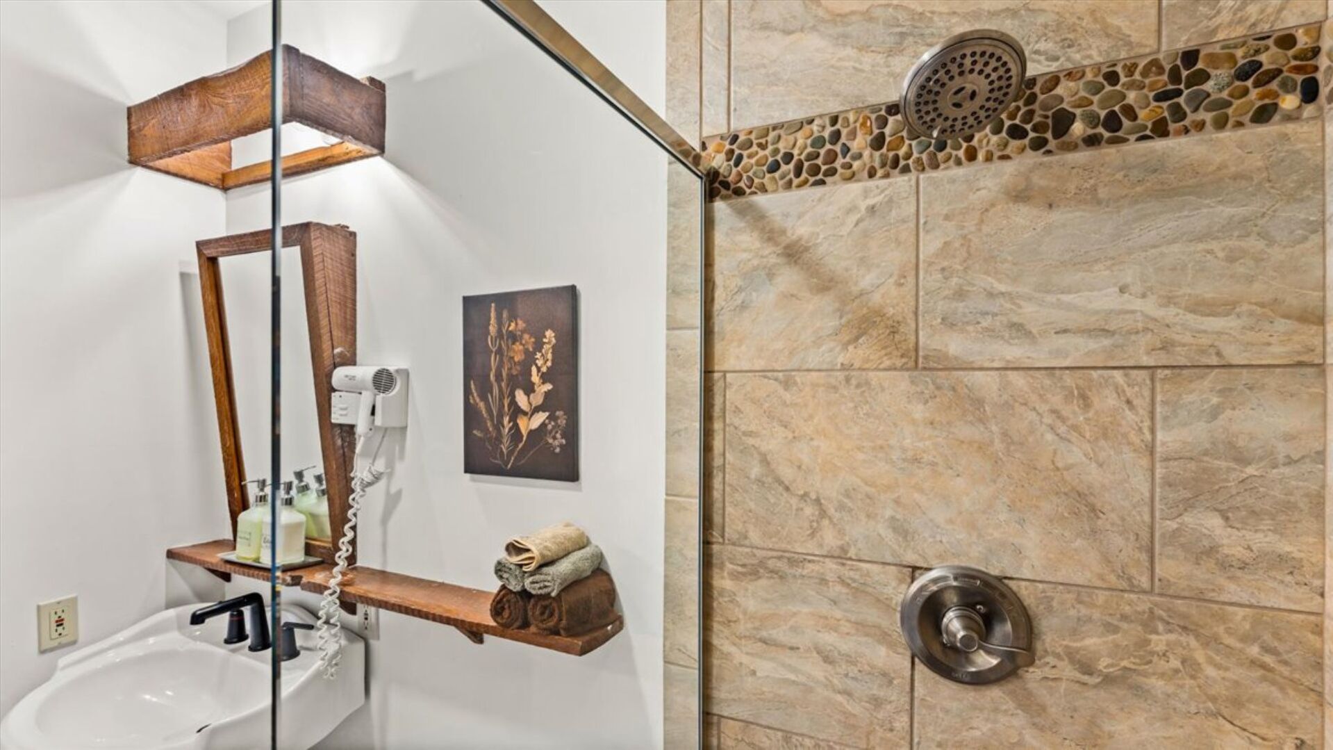 Un baño con un lavabo blanco, un secador de pelo, un estante de madera con toallas, un espejo, un cuadro enmarcado y una ducha con detalles de mosaico de piedra.