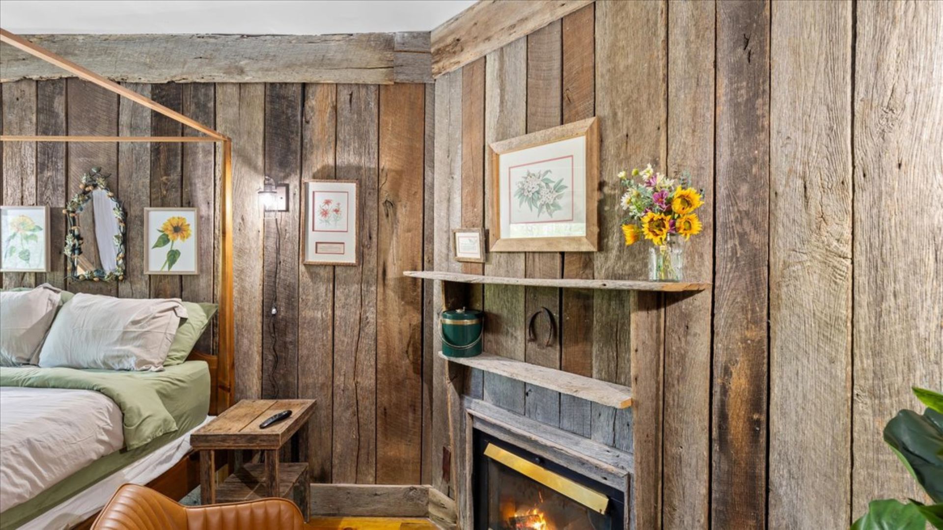 A rustic bedroom with a wooden wall, a bed with green bedding, a small wooden table, a shelf with a vase of flowers, and a fireplace.