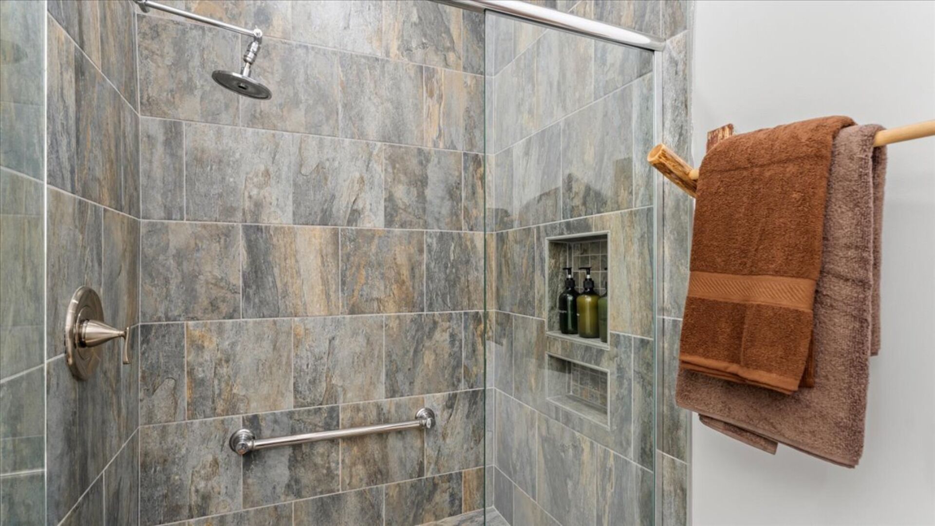 A shower area with a glass door, a handheld showerhead, a towel rack with a brown towel, and a small shelf with toiletries.