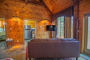 A log cabin interior with a sofa, a television, a kitchen area, and a dining table.