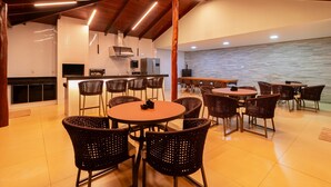 A spacious indoor area with multiple round tables and wicker chairs, a kitchen area with modern appliances, and a view of the water. - Joia Rara Pousada (Pirenópolis)