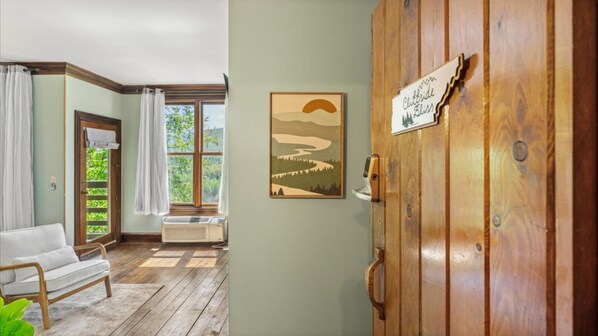 Una habitación con puerta de madera, un cuadro con paisaje enmarco, un sofá blanco y una ventana con cortinas.