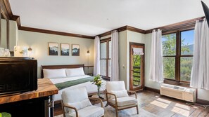 Ein Hotelzimmer mit einem Bett, zwei Sesseln, einem Schreibtisch mit Fernseher und großen Fenstern mit Vorhängen.