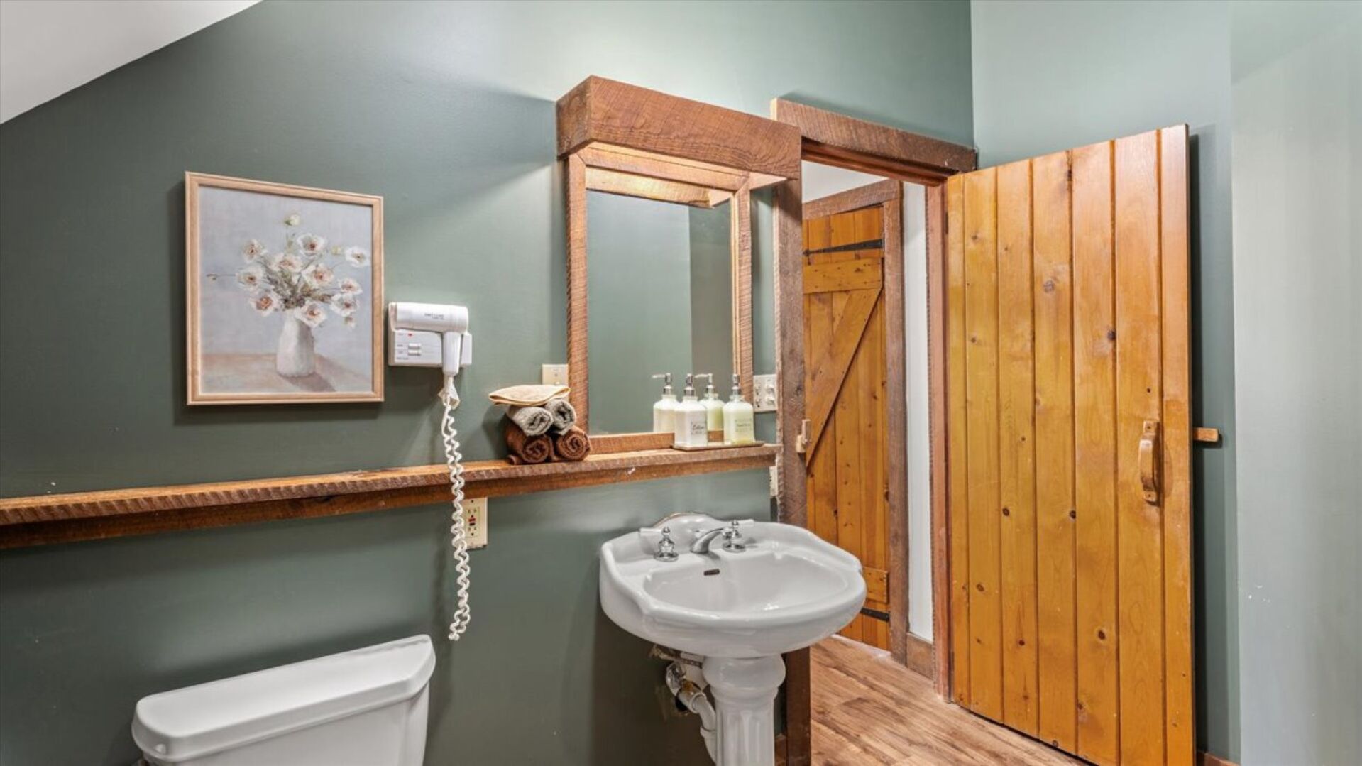 Ein Badezimmer mit einer weißen Toilette, einem Standwaschbecken, einem gerahmten Blumenbild, einem Fön und einer Holztür.