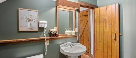 A bathroom with a white toilet, a pedestal sink, a framed floral painting, a hairdryer, and a wooden door.