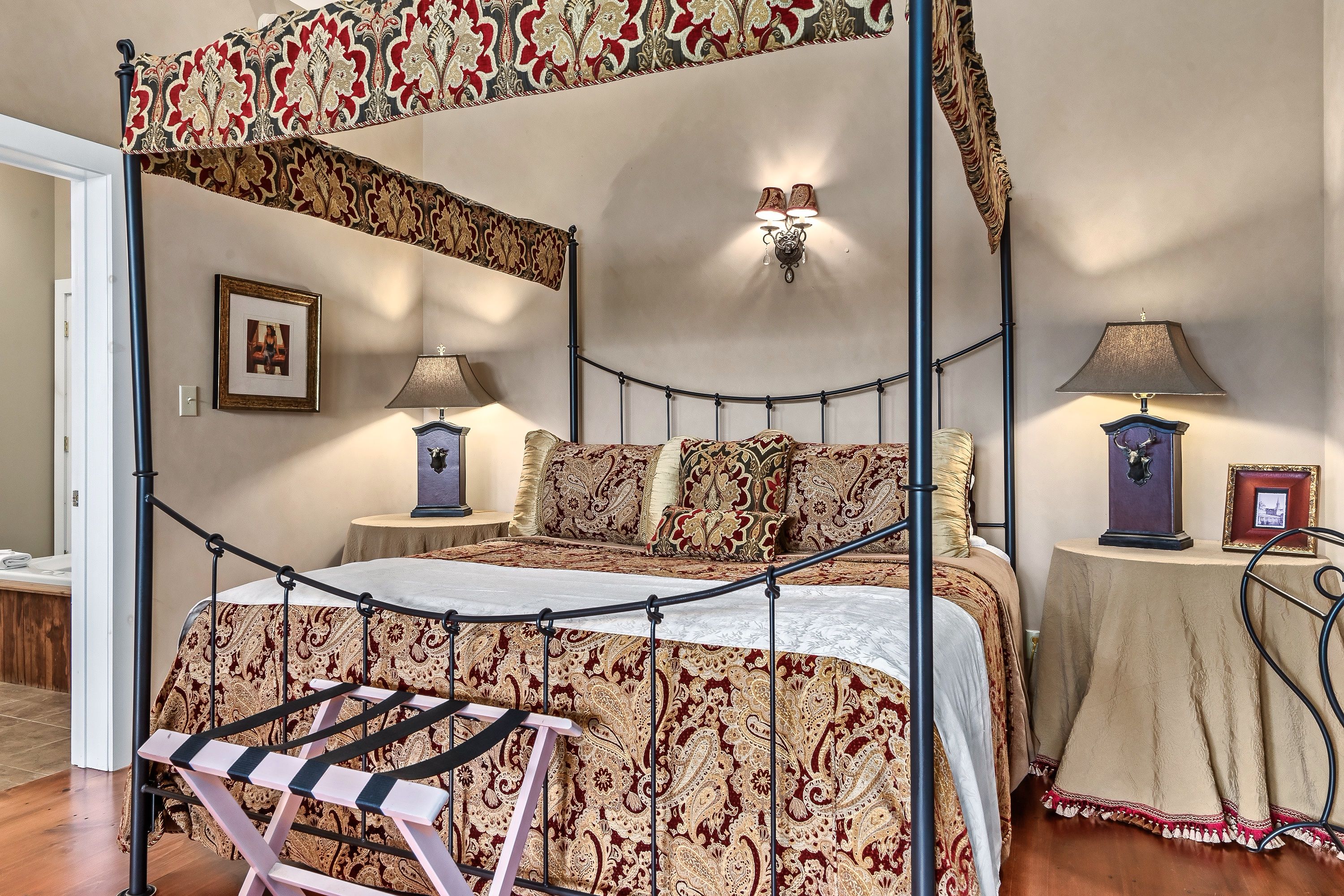 A four-poster canopy bed with a patterned bedspread, two bedside tables with lamps, a framed picture on the wall, and a small table with a lamp and a framed picture.