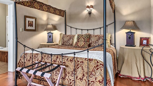 A four-poster canopy bed with a patterned bedspread, two bedside tables with lamps, a framed picture on the wall, and a small table with a lamp and a framed picture.