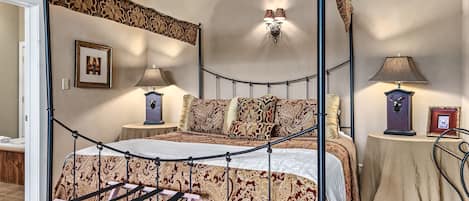 Una cama con dosel y cortinas, un edredón estampado, dos mesitas de noche con lámparas, un cuadro enmarcado en la pared y una pequeña mesa con una lámpara y un cuadro.