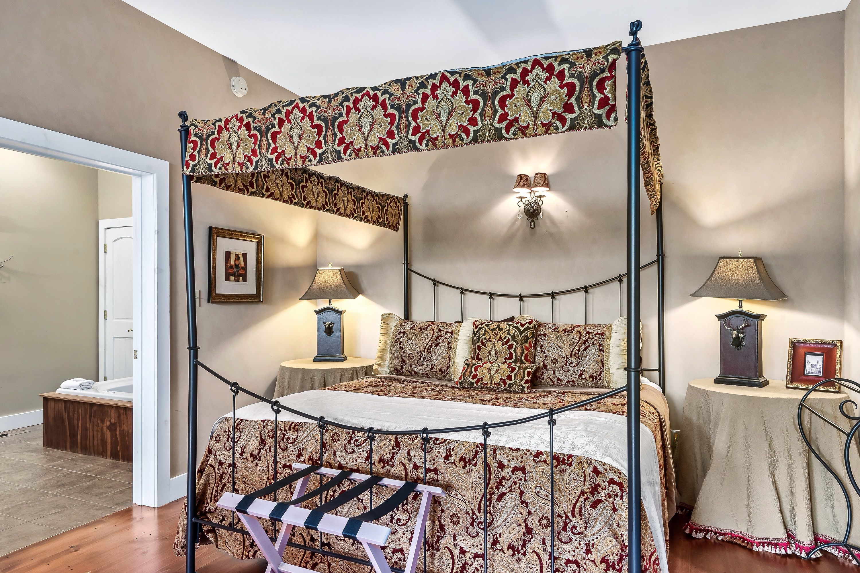A four-poster canopy bed with a patterned bedspread, two bedside lamps, and a small bedside table with a picture frame.