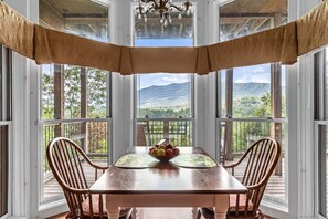 A dining area with a table and chairs, a view of a mountainous landscape through large windows, and a chandelier. - Bordeaux - Mountain View Suite with Whirlpool Tub & Fireplace (Townsend)