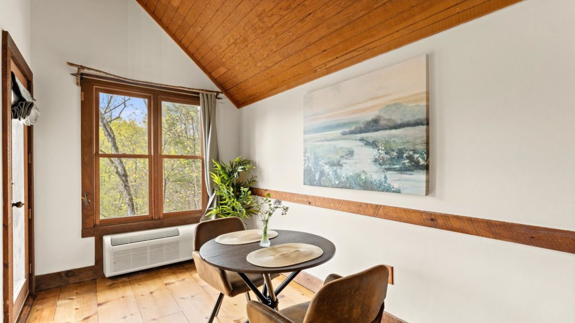 A dining area with a table and chairs, a window with a view of trees, a painting on the wall, and a wooden ceiling.
