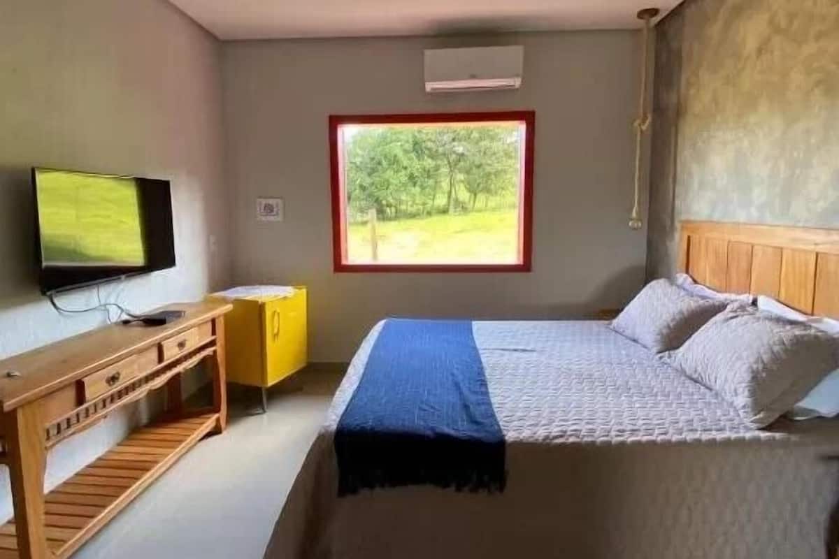 A bedroom with a bed, a TV, a desk, and a window with a view of trees.