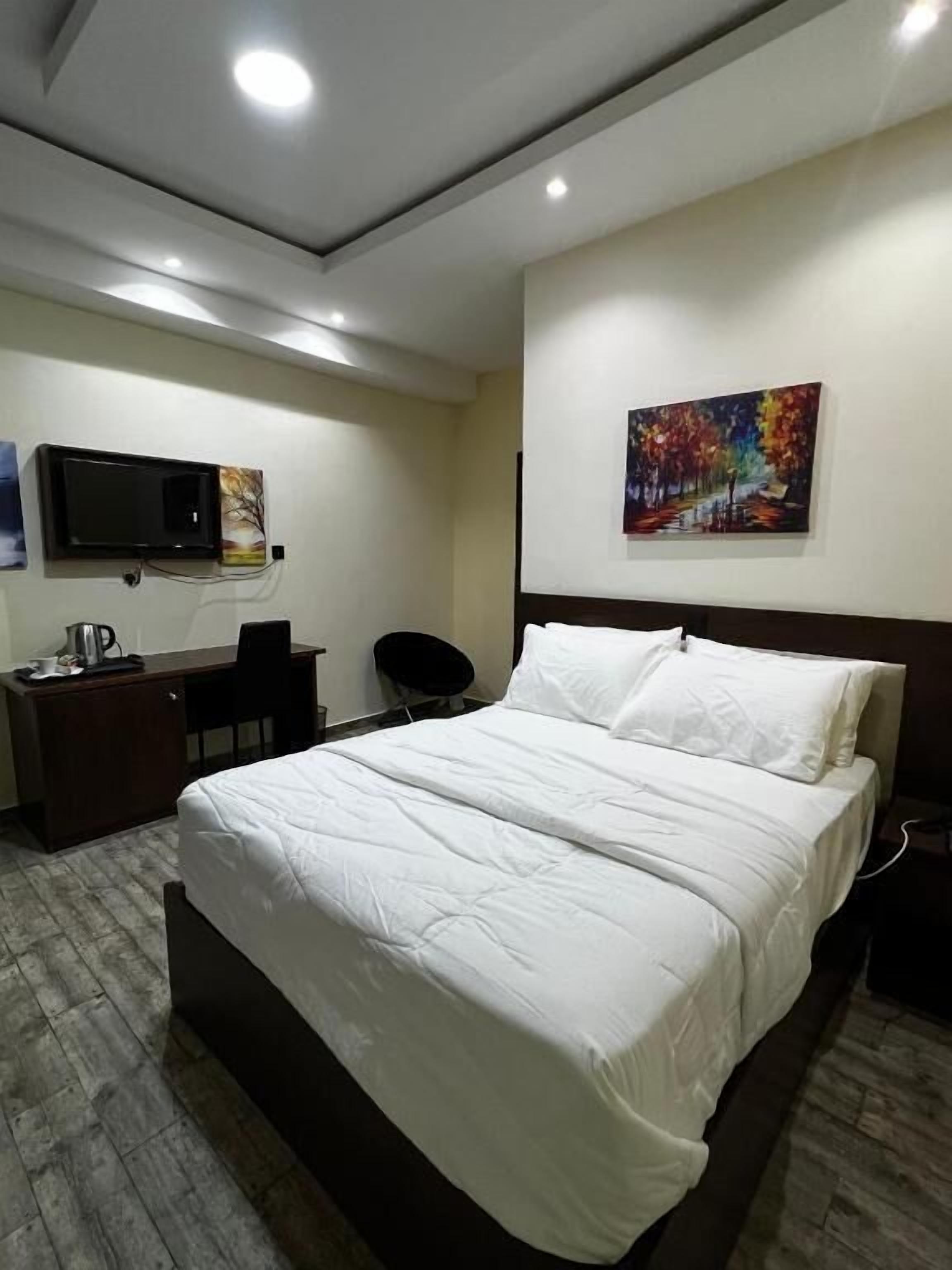 A hotel room with a bed, a desk, a chair, a TV mounted on the wall, and a painting.