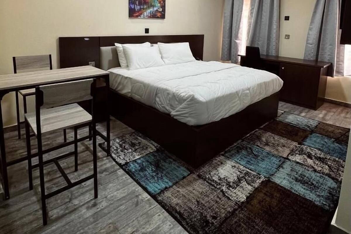 A modern bedroom with a large bed, a desk, and a chair. There is a painting on the wall, a window with curtains, and an air conditioning unit.