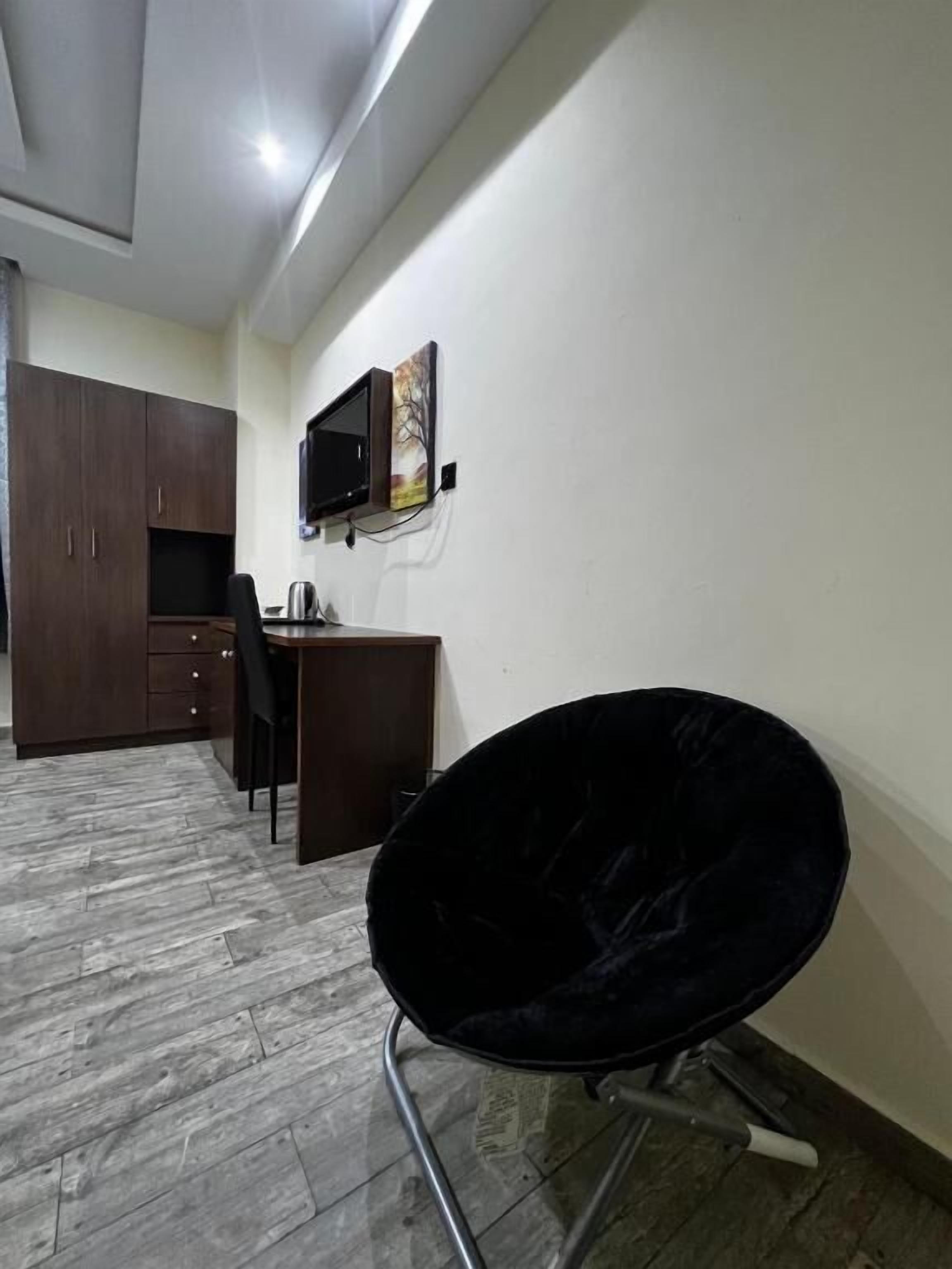 A room with a dark wooden wardrobe, a desk with a chair, a mounted TV, and a black chair.