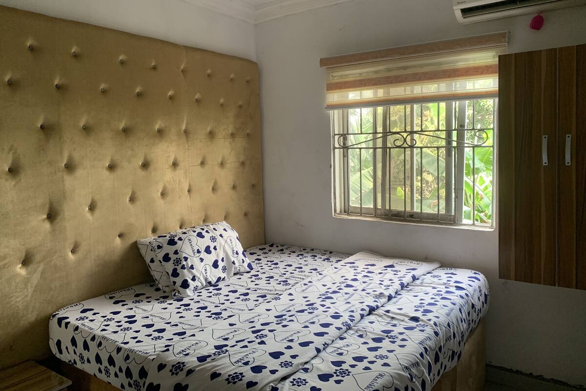 A bedroom with a tufted headboard, a bed with patterned bedding, a window with metal bars, and a wardrobe.