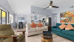 Una sala de estar moderna con sofá, sillón y una mesita de centro, con una cocina al fondo y un ventilador de techo.