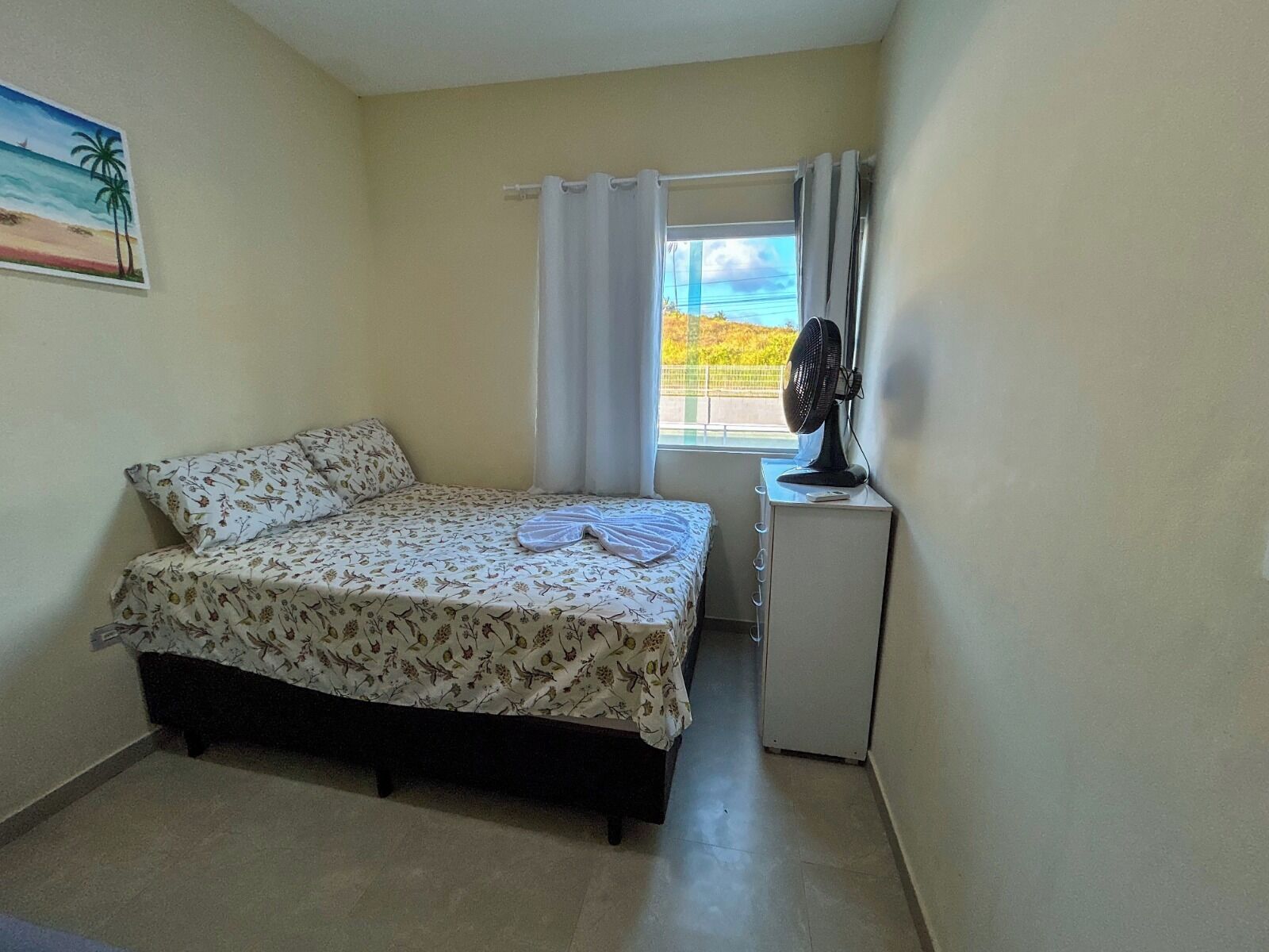 A small bedroom with a bed, a desk, a fan, and a window with curtains.