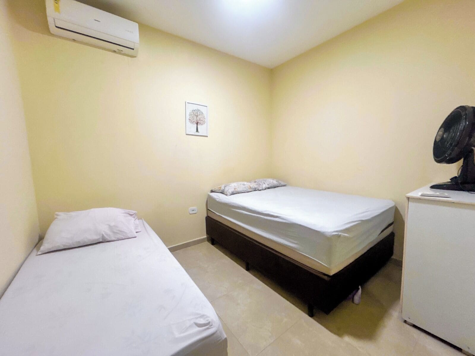 A small, clean room with two single beds, a white desk, a black fan, and an air conditioner on the wall.