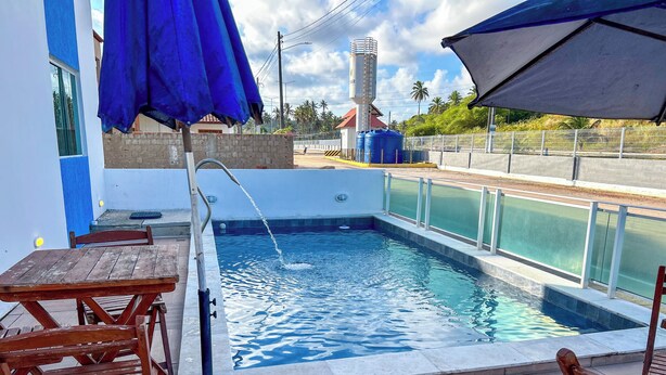 A swimming pool with a water fountain, surrounded by wooden chairs and a blue umbrella. - Flats Praia de Gravatá (São José da Coroa Grande)