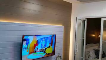 Uma sala de estar moderna com uma televisão de tela plana na parede, um armário branco com diversos objetos, um relógio e uma porta de vidro deslizante que dá para um quarto.