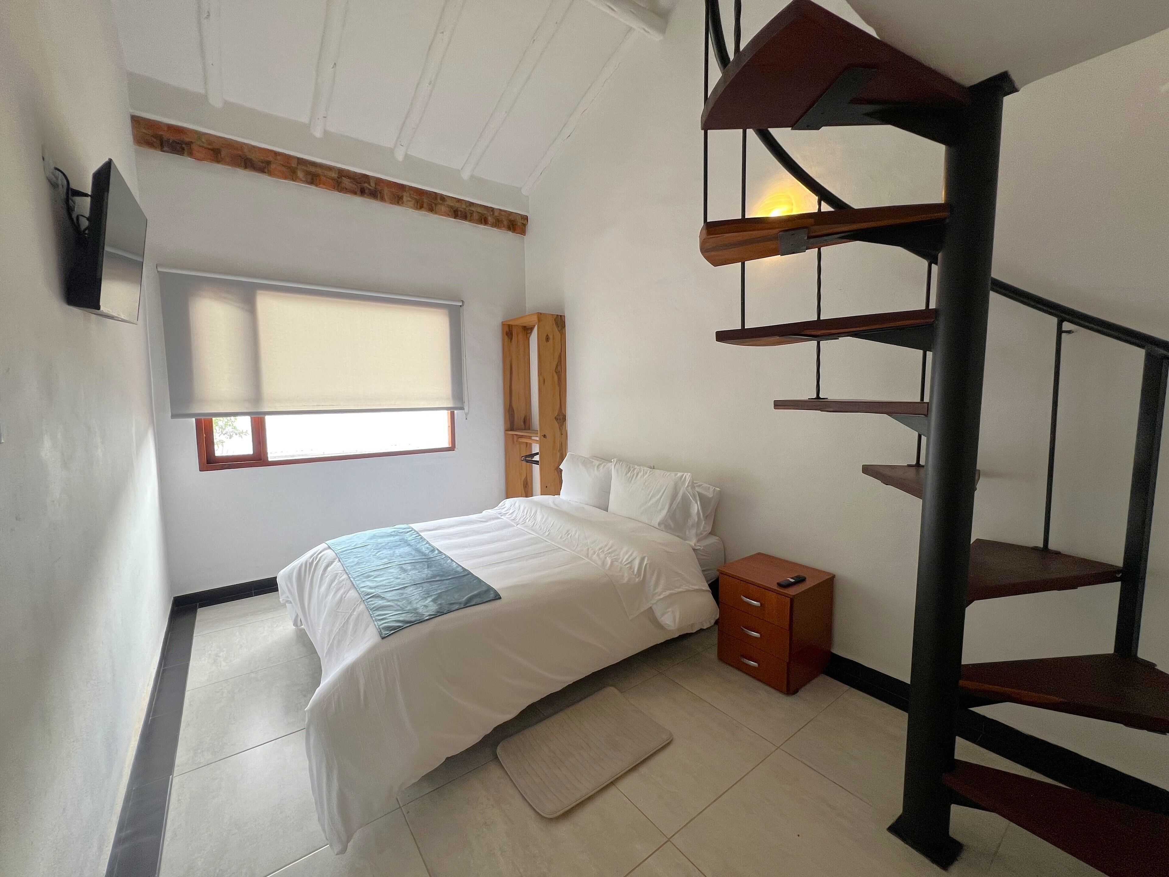A bedroom with a bed, a nightstand, a wooden wardrobe, a window with blinds, and a spiral staircase.